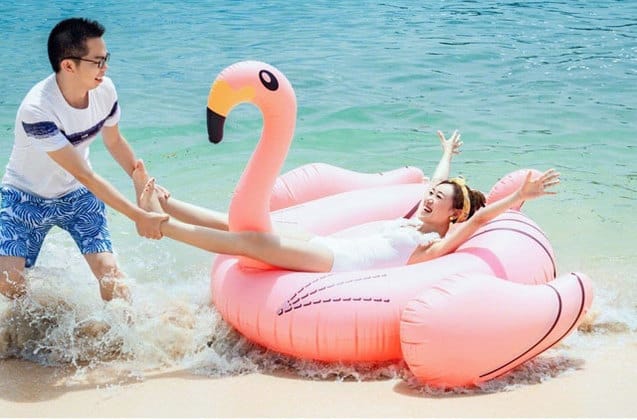刁曼岛海滩上男子拉着躺在水上漂浮充气火烈鸟上笑着的女朋友的脚