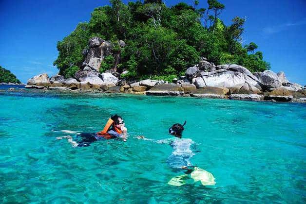 two people are snorkeling at crystal clear green seawater in redang island with beautiful view of rocks and tress in the background