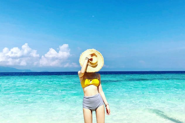 woman in yellow bikini standing at island beach holding a straw hat blocking her face