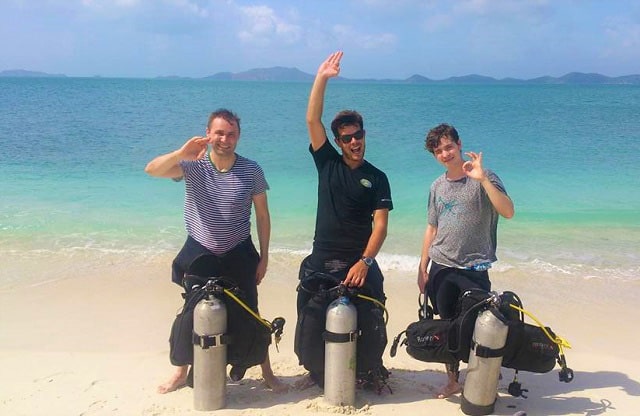 three men are waving on the beach of sibu island with diving equipment in front of simba resort