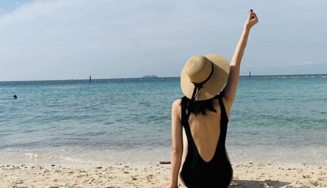 woman in black swim suit and straw hat sitting on koh lipe island beach facing sea with one arm raised