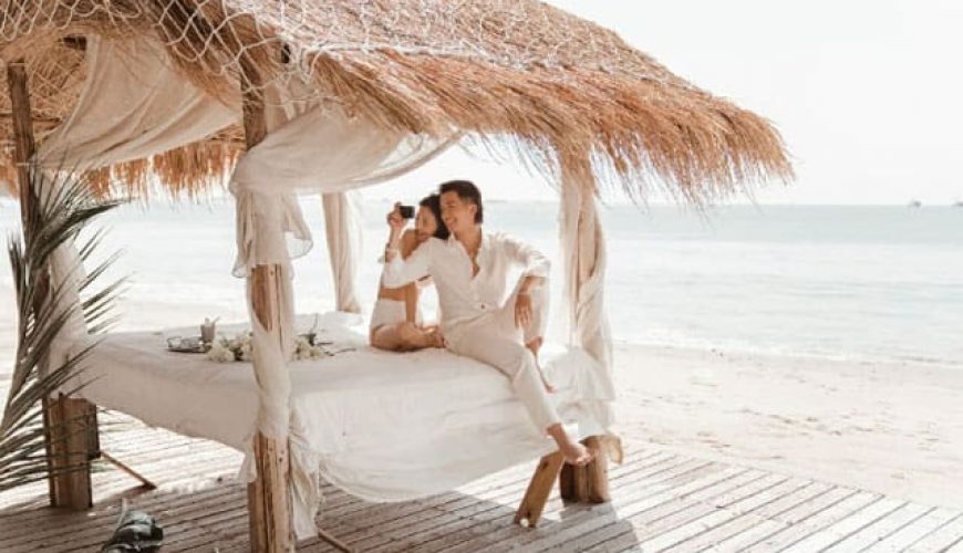 couple in white clothes sitting on bed below big straw umbrella in tenggol island